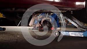 woman mechanic with tattoos lies under car on car repair trolley and repairs it with tools. View of girl in blue