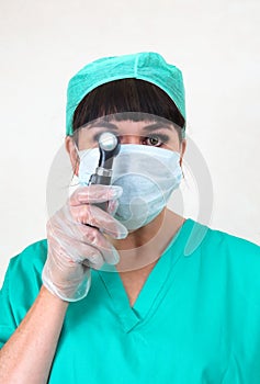 Woman medic in face mask holding otoscope to eye.