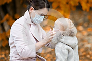 Woman in a medical face mask adjusts the medical mask to baby girl, being in the yard in the fall
