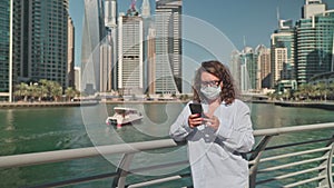 Woman in medical face mask browsing phone walking on waterfront in city.