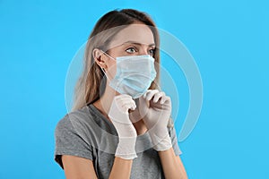 Woman in medical gloves putting on protective face mask against light blue background