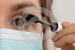 Woman in a medical mask does makeup, mascaras her eyes. The face of a young beautiful woman in a stylish mask