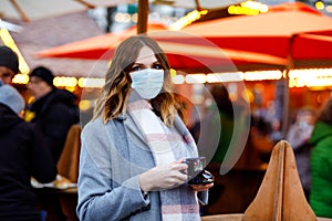 Woman with medical mask drinking hot punch, mulled wine on German Christmas market. People with masks as protection