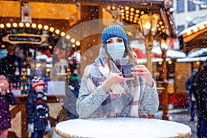 Woman with medical mask drinking hot punch, mulled wine on German Christmas market. People with masks as protection