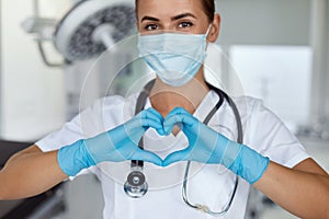 Woman with a medical mask and hands in latex glove shows the symbol of the heart. Doctor for the heart.