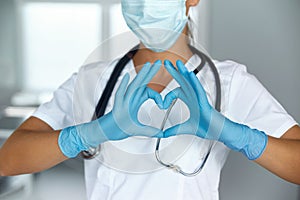 Woman with a medical mask and hands in latex glove shows the symbol of the heart. Doctor for the heart