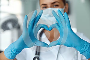 Woman with a medical mask and hands in latex glove shows the symbol of the heart. Doctor for the heart.