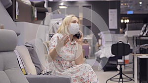 A woman in a medical mask on her face is sitting in a shopping center talking on the phone