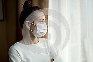 A woman in a medical mask looks at the street through a curtained window