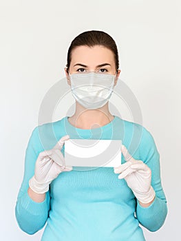 A woman in a medical mask and surgical gloves holds a small white tablet with place for text. Shot on a light background.