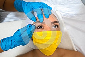 Woman in a medical mask waxing her eyebrow, up close. Woman in a beauty parlor. Plastic surgery clinic