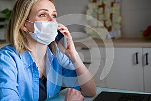Woman in medical mask is working office work remotely from home on kitchen. Talking mobile phone. Using computer