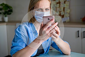 Woman in medical mask is working office work remotely from home on kitchen. Talking mobile phone. Using computer