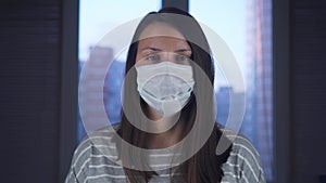 A woman in a medical protective mask stands at home against the background of the city outside the window. The concept