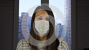 A woman in a medical protective mask stands at home against the background of the city outside the window. The concept