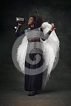 Woman, medieval warrior with wings and chainmail holding vintage video camera and binoculars against dark background