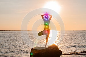 Woman is meditating with glowing seven chakras on the beach. Silhouette of woman is practicing yoga at sunset