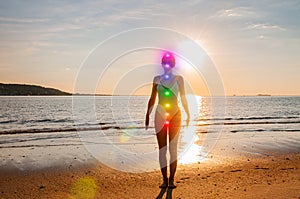 Woman is meditating with glowing seven chakras on the beach. Silhouette of woman is practicing yoga at sunset