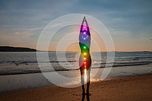 Woman is meditating with glowing seven chakras on the beach. Silhouette of woman is practicing yoga at sunset