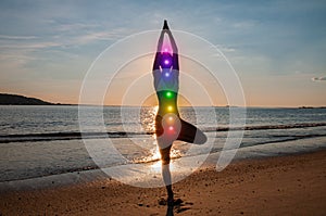 Woman is meditating with glowing seven chakras on the beach. Silhouette of woman is practicing yoga at sunset