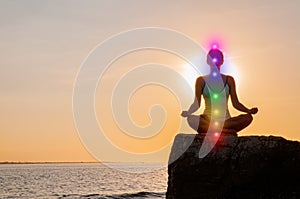 Woman is meditating with glowing seven chakras on stone at sunset. Silhouette of woman is practicing yoga on the beach