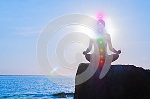 Woman is meditating with glowing seven chakras on stone at sunset. Silhouette of woman is practicing yoga on the beach