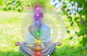 Woman is meditating in the lotus pose with glowing seven chakras on grass. Woman is practicing yoga on the park