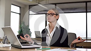 Woman meditating at office desk, reducing work stress and irritation, relaxing