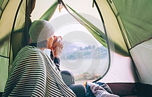 Woman meets cold morning sitting in touristic tent with cup of hot tea. Active vacation concept image.