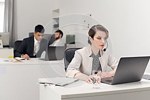 Woman with a micro sit at a table with laptop, collegues are busy with work