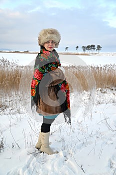 The woman of middle age in a fur cap and a colorful shawl costs on the bank of the winter lake