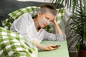A woman of middle age, sporting a short haircut, stays in bed all day, fixated on her phone. Indolence, apathy, seeking photo