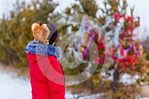 A woman in a mink and red down jacket stands with her back to the camera and looks at a Christmas tree in the woods in winter