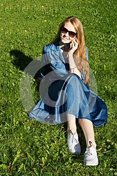 Woman and mobile phone, green lawn, summer. Red hair girl, blue dress, sitting on the grass outside, holding a telephone.