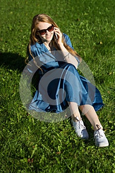 Woman and mobile phone, green lawn, summer. Red hair girl, blue dress, sitting on the grass outside, holding a telephone.