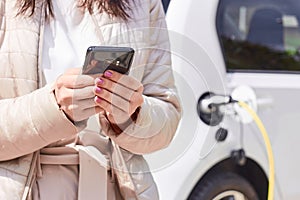 Woman with a mobile phone near recharging electric car. Vehicle charging at public charging station outdoors. Car sharing concept