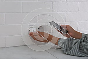 Woman with mobile phone plugging charger in socket on white brick wall, closeup