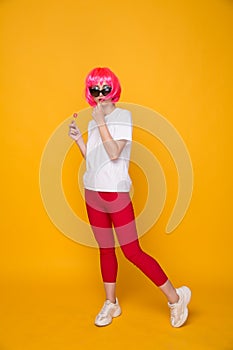 Woman model in the yellow studio with lollipop in a pink wig and bright clothes.