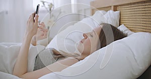 Woman in morning sitting in bedroom leaning against the soft leather back of bed. Write messages with your smartphone