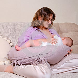 Woman mother breastfeeds an infant baby sitting on a home sofa. Mom feeds milk toddler baby in living room