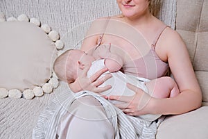 Woman mother breastfeeds an infant baby sitting on a home sofa. Mom feeds milk toddler baby in living room