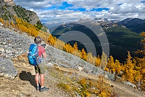 Woman on a mountain top looking at scenic view of mountains and yellow larch trees from above.