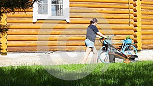 Woman mowing green lawn in backyard with lawn mower moving along wooden country house on summer sunny day. Landlady