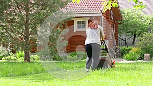 Woman mowing green lawn in backyard near wooden country house moving away area with lawn mower on summer sunny day back