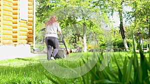 Woman mowing green lawn in backyard near wooden country house moving away area with lawn mower on summer sunny day back