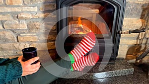 Woman with a mug sits near a hot burning fireplace, legs stretched out in red striped socks to the fire, winter relaxation concept