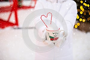 Woman with mug with snow, candy cane and inscription Merry and Bright in her hands outdoor in warm clothes in winter festive