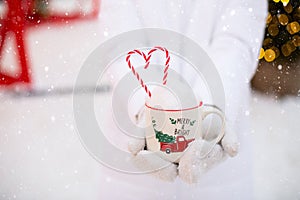 Woman with mug with snow, candy cane and inscription Merry and Bright in her hands outdoor in warm clothes in winter festive