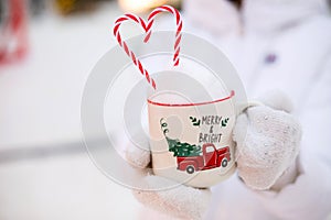 Woman with mug with snow, candy cane and inscription Merry and Bright in her hands outdoor in warm clothes in winter festive