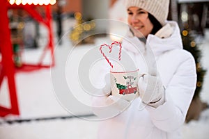 Woman with mug with snow, candy cane and inscription Merry and Bright in her hands outdoor in warm clothes in winter festive